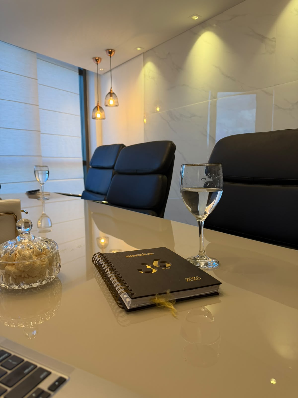 Sala do escritório com mesa de reunião, cadeiras pretas e luminárias de luz quente.