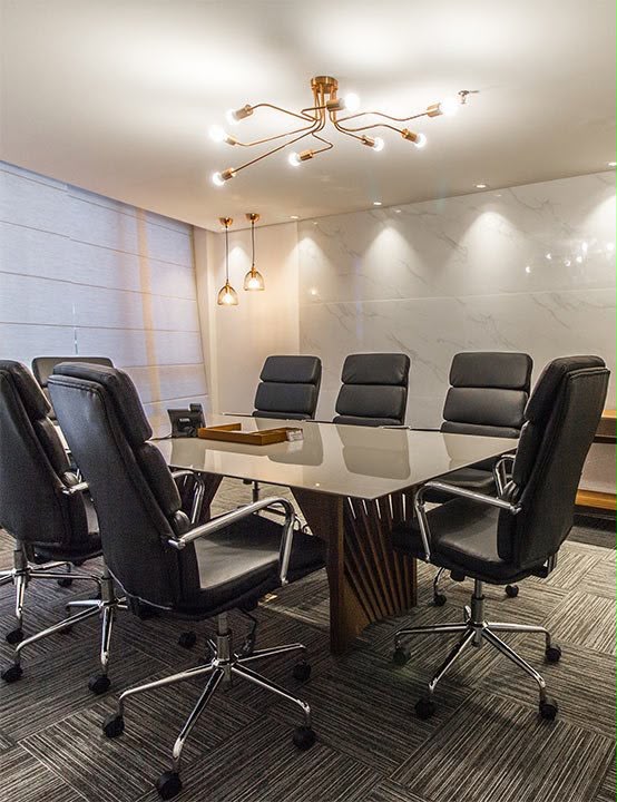 Sala de reunião com mesa retangular, cadeiras pretas, parede de mármore claro e luminárias douradas.